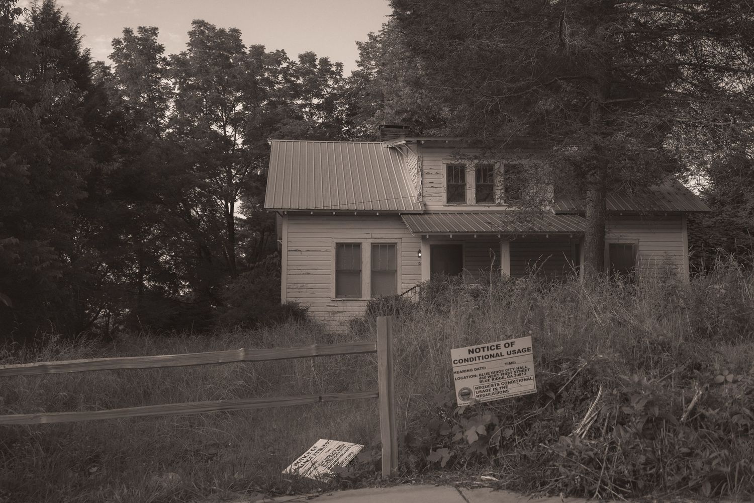 Abandoned vintage house with overgrown yard, metal roof, and warning sign in sepia-toned black and white photo.