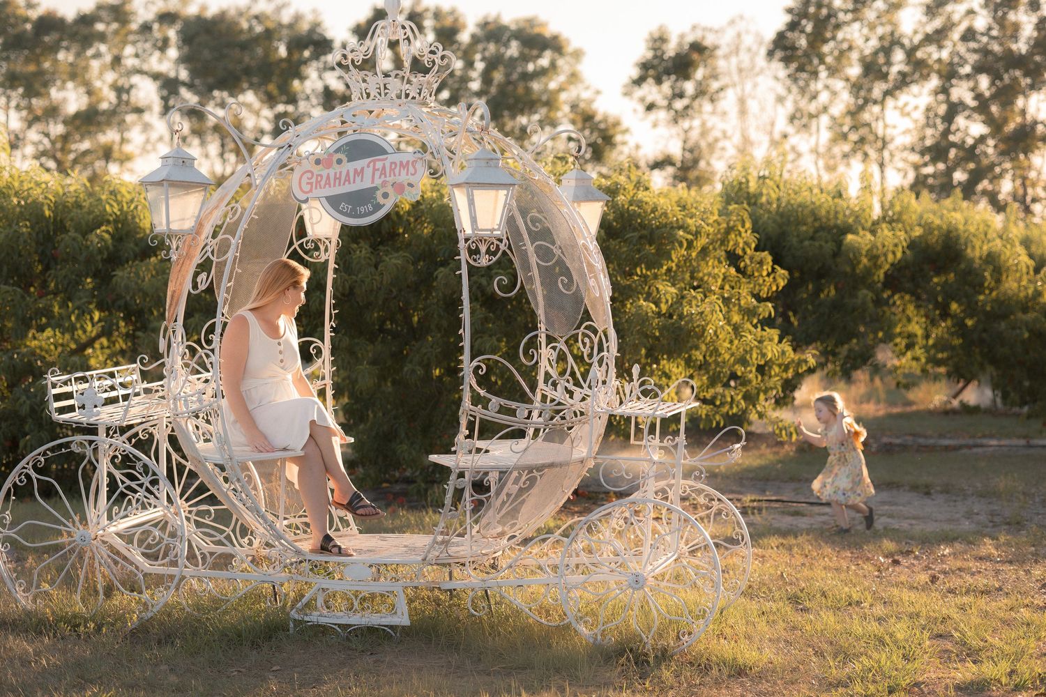 Woman in white dress sits in ornate white Cinderella carriage outdoors while child plays nearby in golden sunlight.