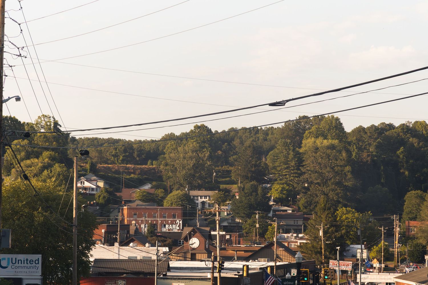 Aerial view of a small town with brick buildings, lush green trees, and utility lines in a hilly landscape.