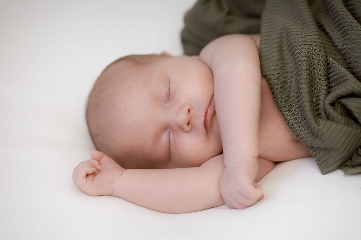 A sleeping baby in olive green swaddle cloth lies peacefully on a white surface.