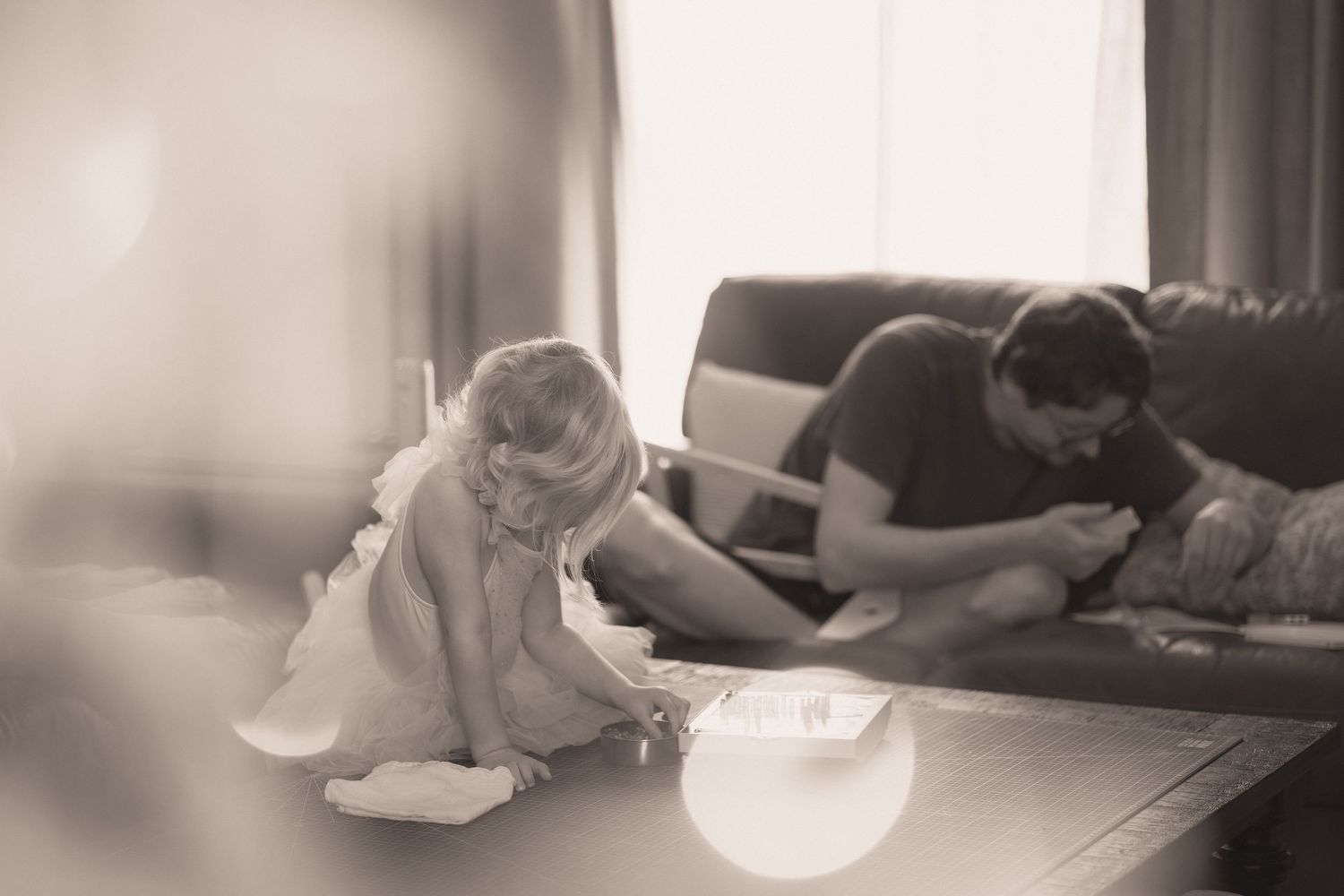 Black and white photo of a child sitting on floor playing while someone rests on couch in background.
