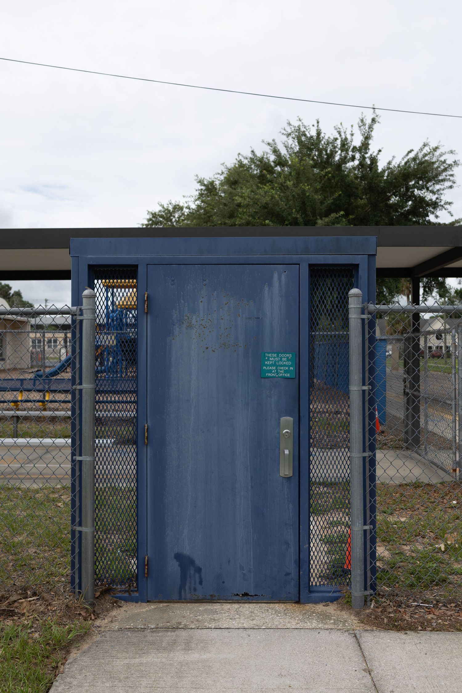 Blue metal security gate with chain-link fence panels at a fenced outdoor facility with trees and structures in background.