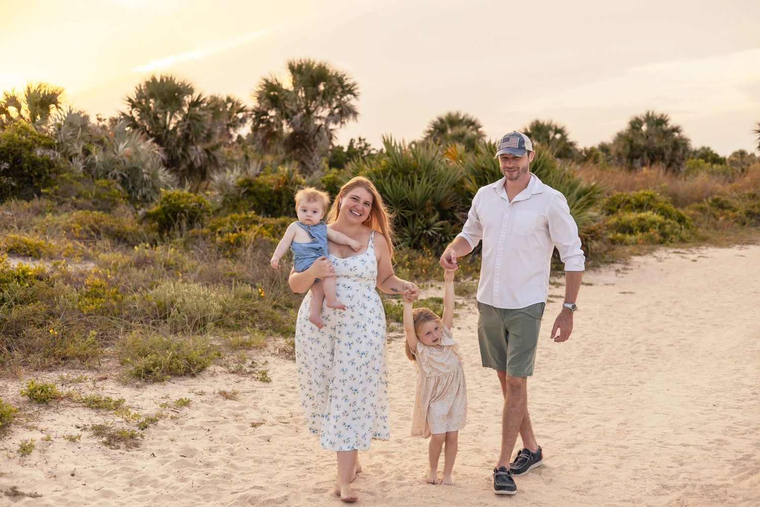 A family walks together on a sandy beach path at sunset with palm trees and coastal vegetation in the background.