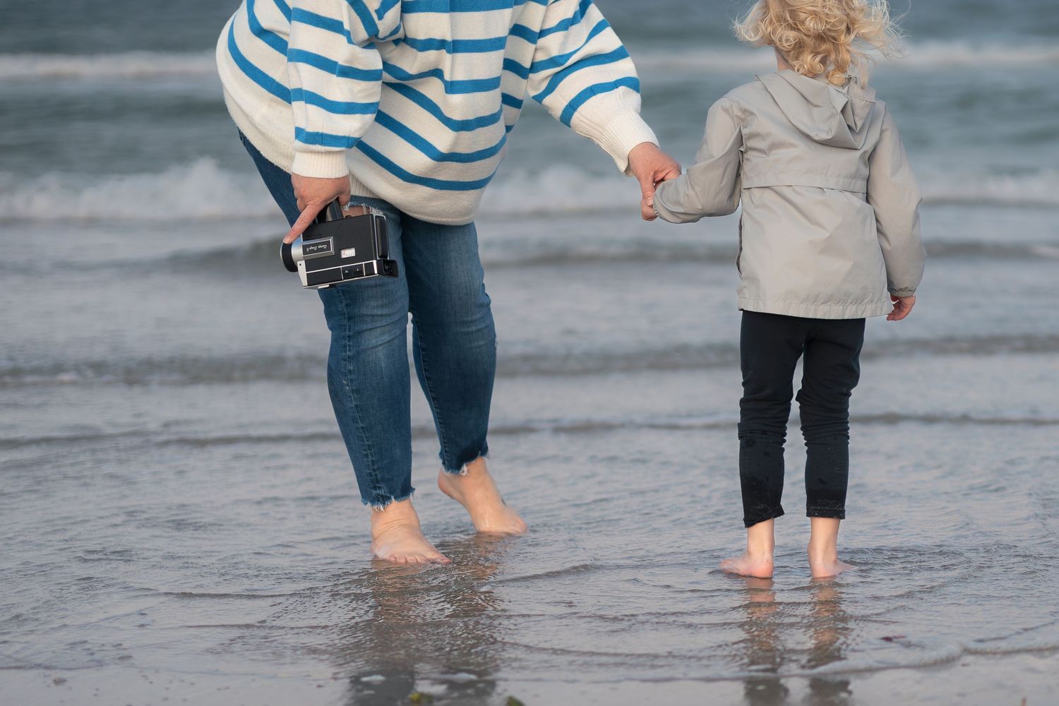 A couple in striped shirts and jeans walking barefoot together along the beach at the water's edge.