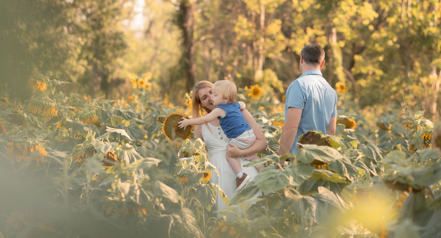 Family with young child sharing a tender moment in a sunflower field surrounded by lush green trees.