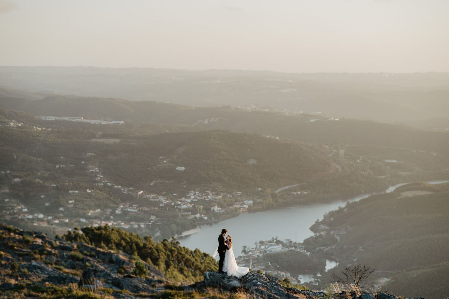 Two silhouettes stand on a mountain overlook with sweeping views of a valley and lake below at sunset.