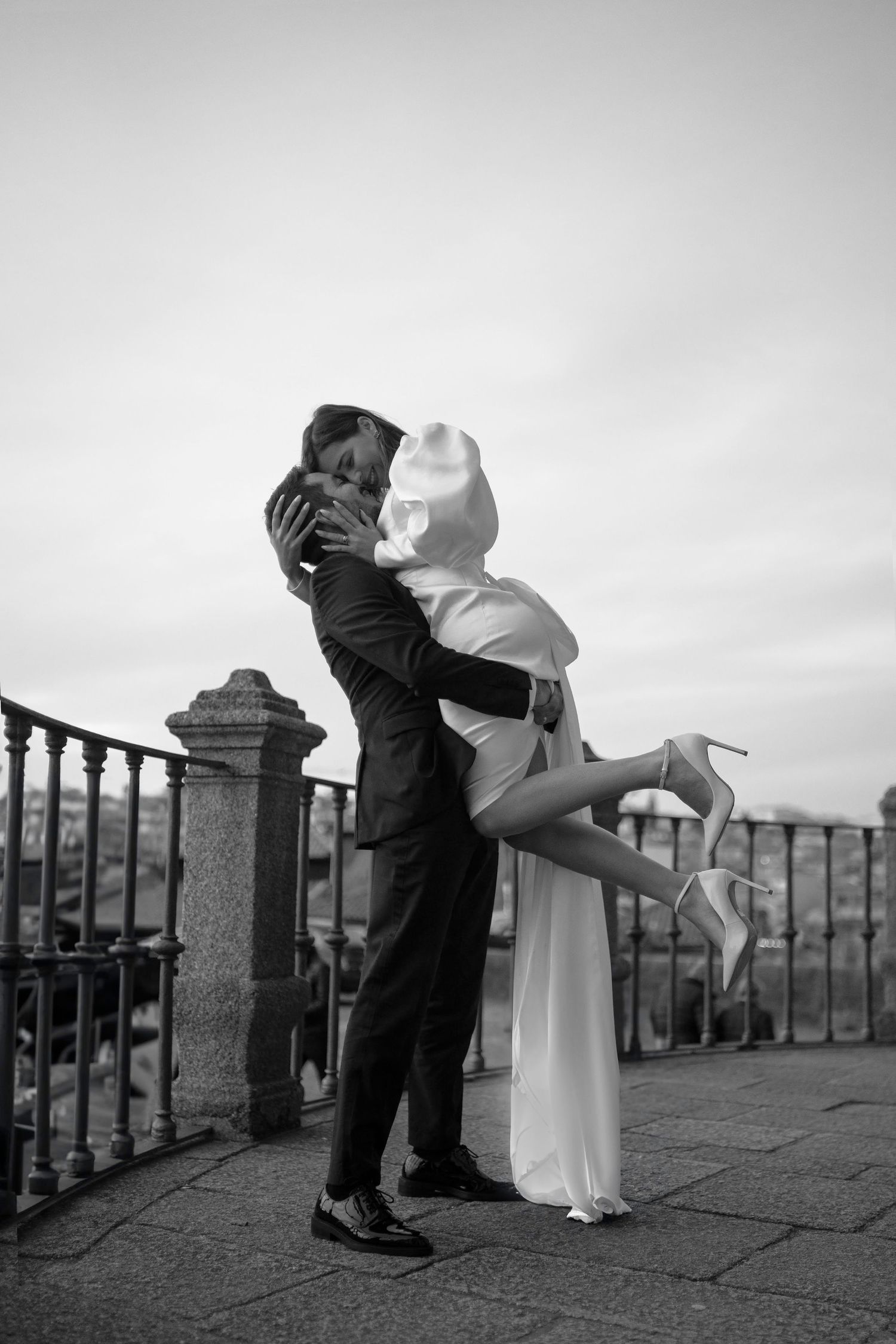 Romantic black and white photo of couple embracing on balcony with bride's leg lifted in classic pose.