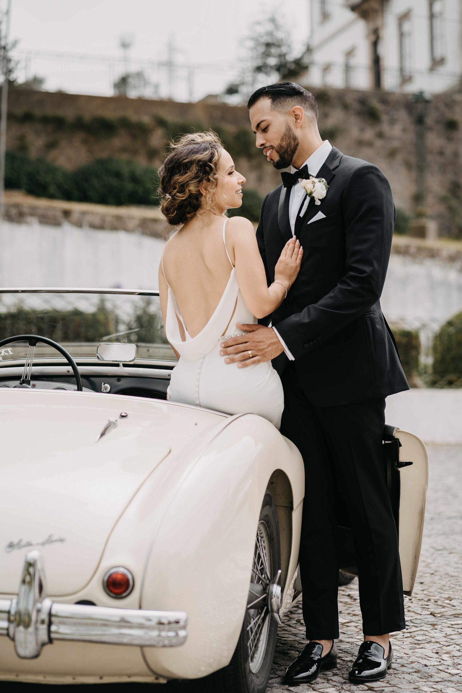 Vintage car wedding photo with couple in elegant attire against stone wall backdrop.