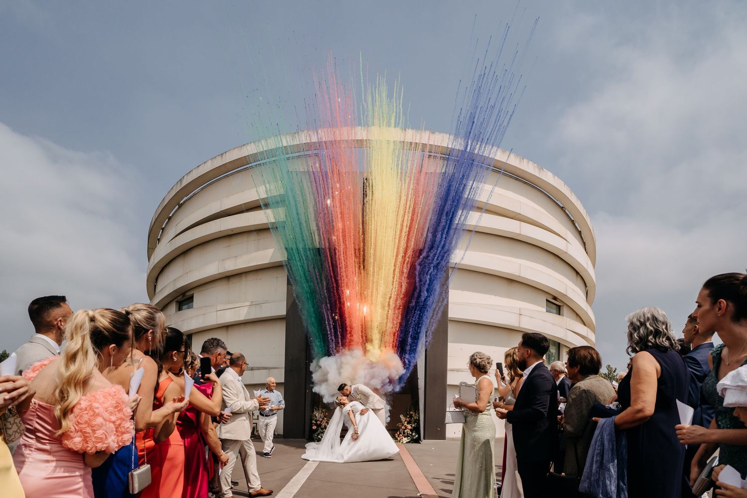 Colorful rainbow smoke effect bursts behind wedding guests gathered on a modern circular building rooftop.