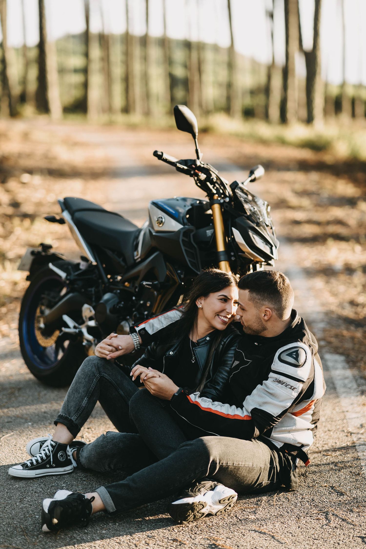 A couple in motorcycle gear share a romantic moment lying on the ground next to their parked sport bike.
