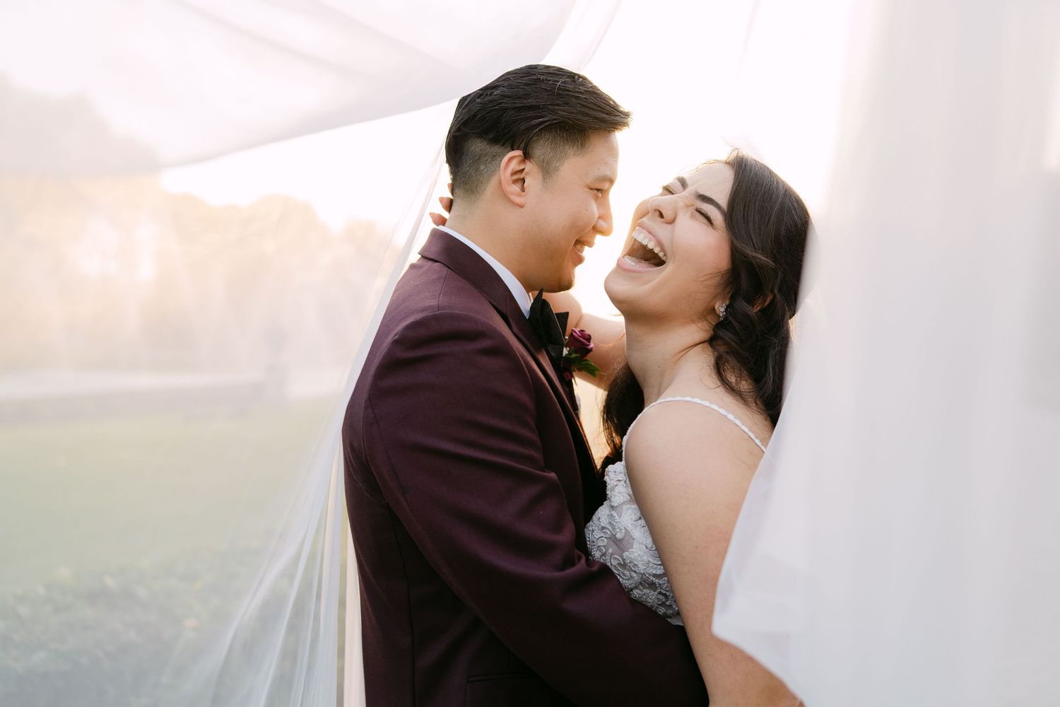 A joyful couple shares an intimate moment in their wedding attire against a misty outdoor backdrop at sunset.