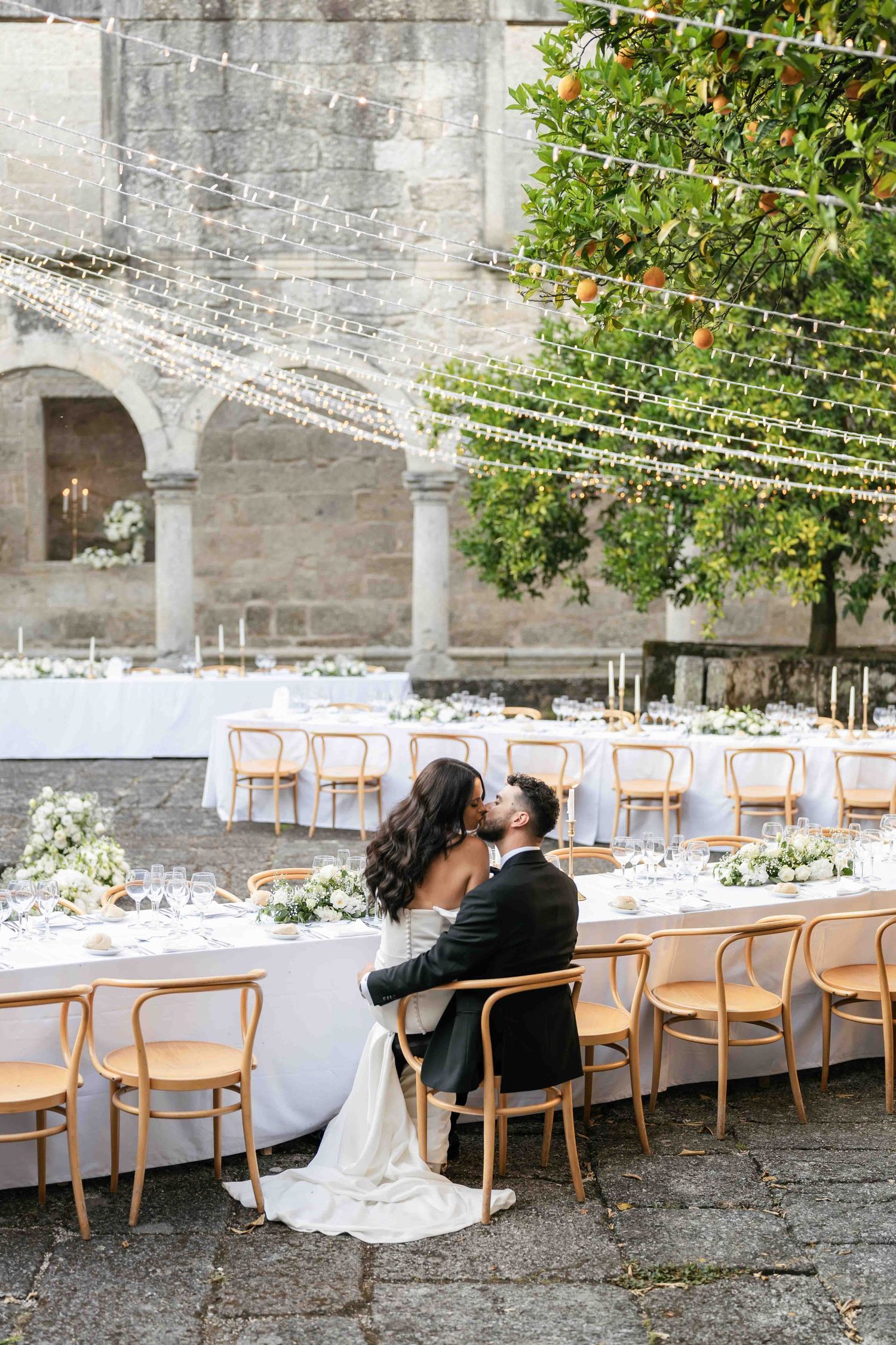 Elegant outdoor wedding reception setup with long tables and wooden chairs under string lights at a historic venue.