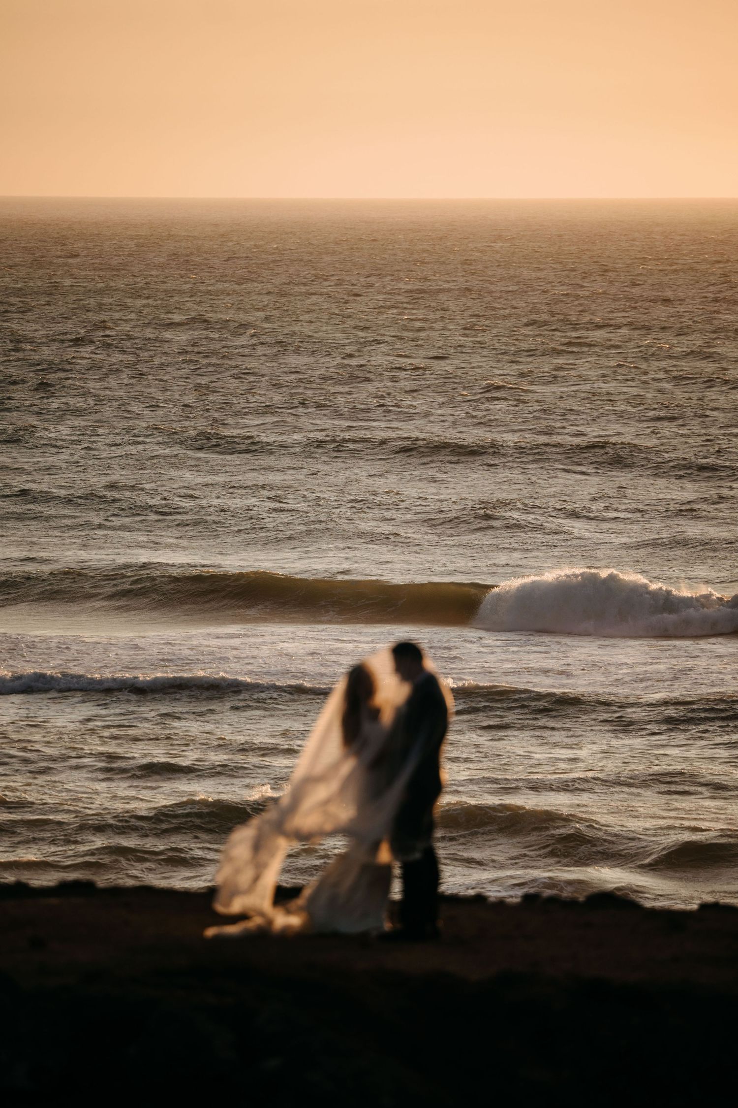 Silhouettes of a couple sitting on a beach cliff at sunset with waves crashing against the shore in the background.
