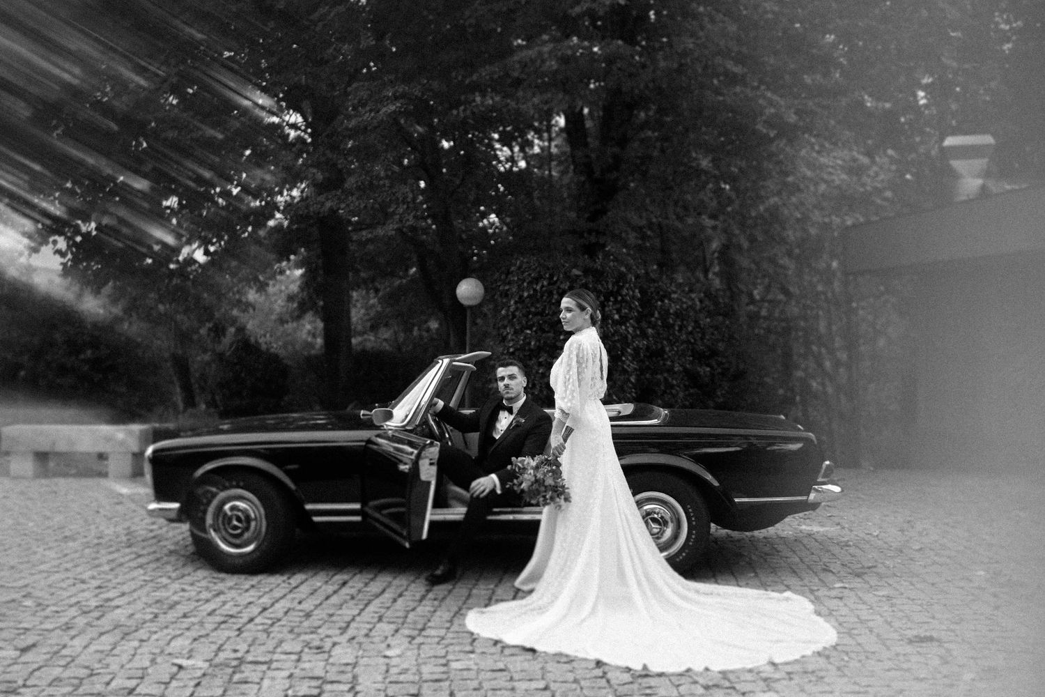 Wedding couple poses beside vintage black car on cobblestone street.