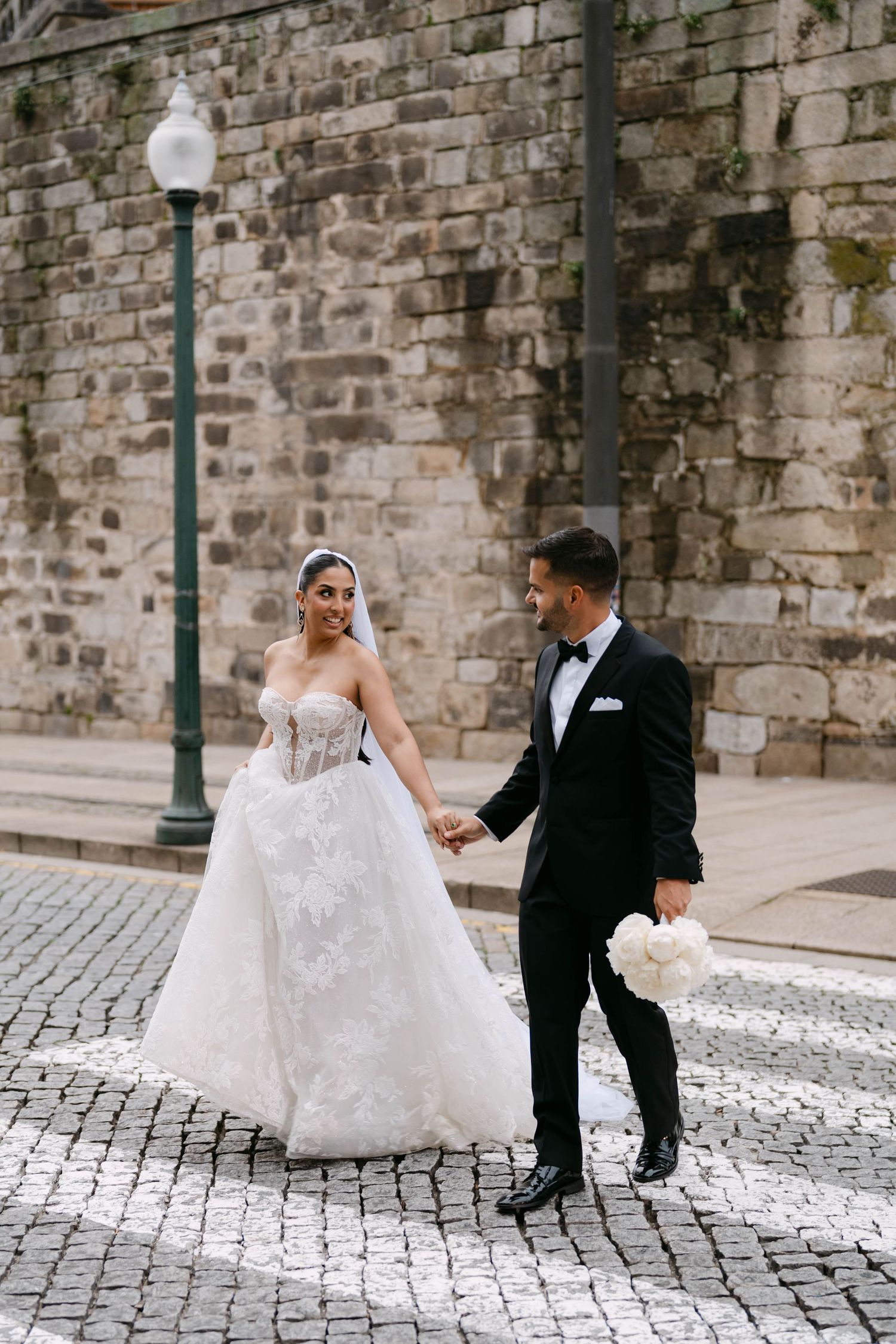 A wedding couple walks along a cobblestone street with a stone wall backdrop.
