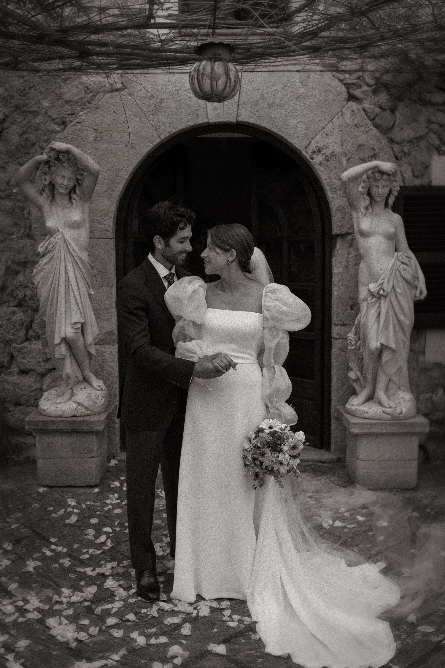 Black and white wedding portrait of couple under archway with classical statues framing the entrance.