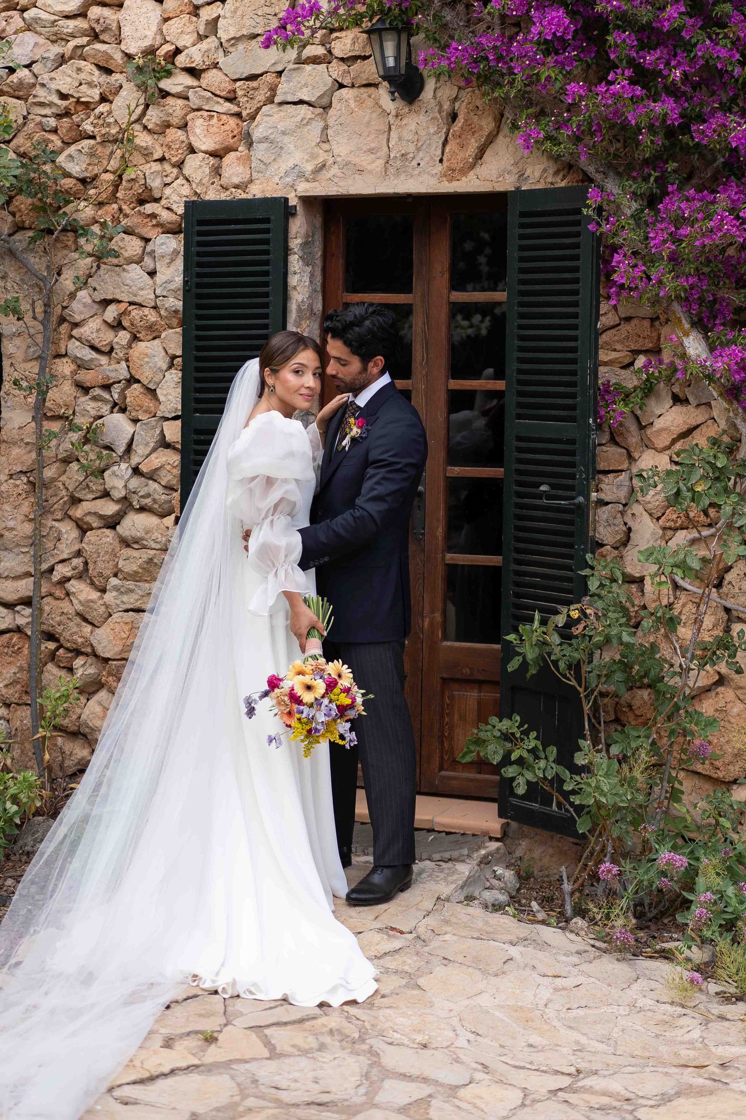 Romantic wedding scene against rustic stone wall with vibrant purple bougainvillea flowers.