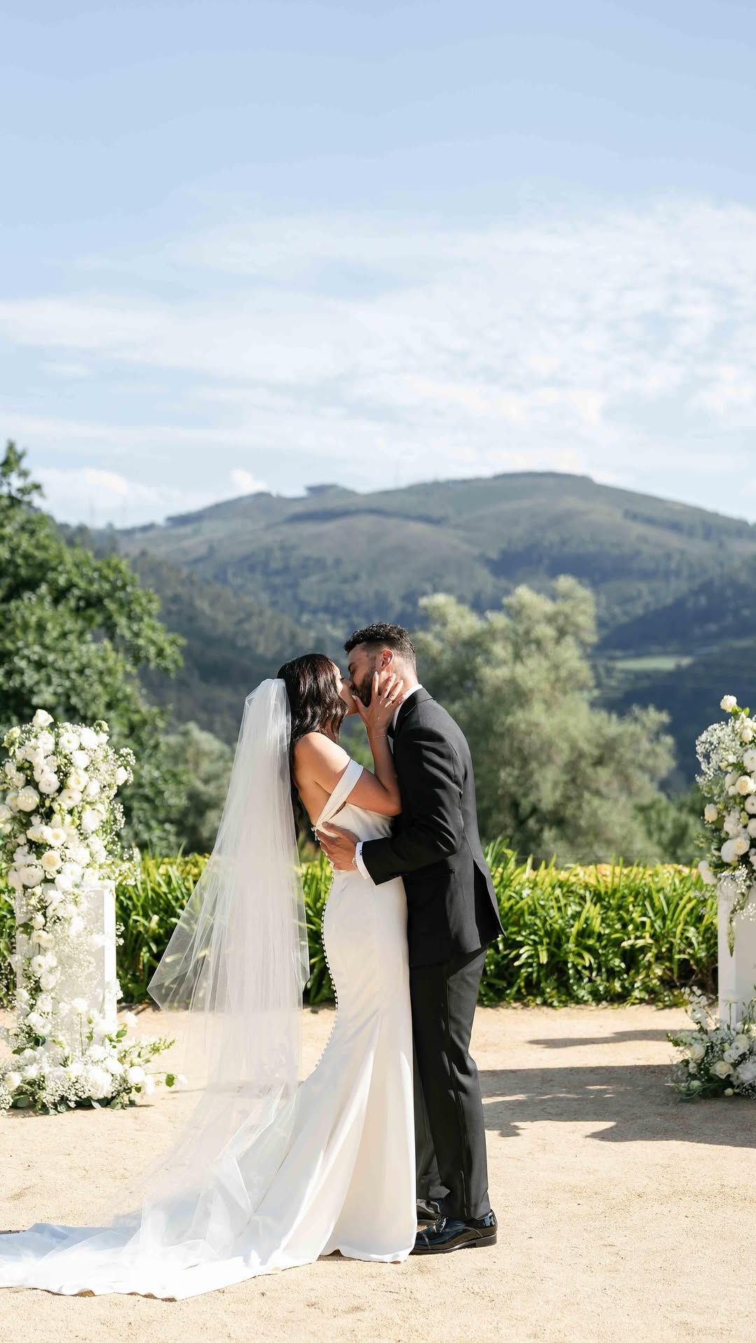 A romantic wedding kiss captured at a scenic vineyard with mountains and greenery in the background.