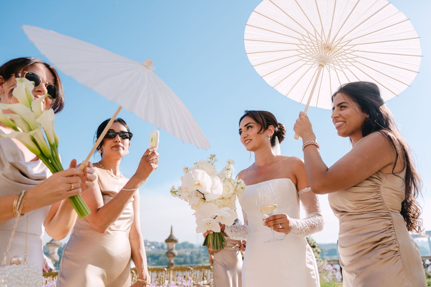 Wedding party members hold white parasols while standing together on a sunny beach during a tropical celebration.