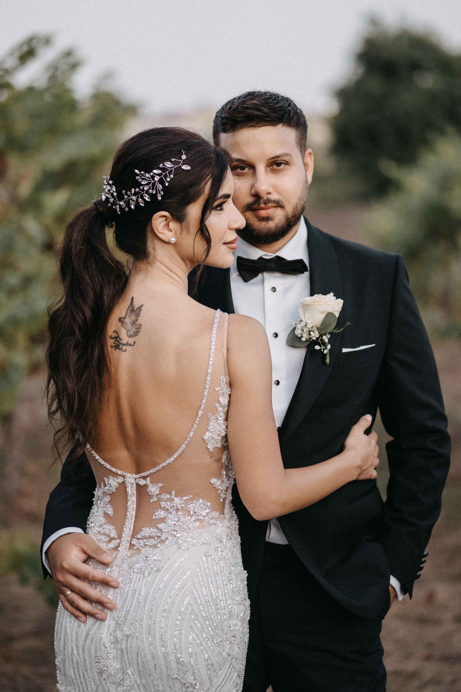 Elegant bride in beaded backless gown embraces groom in black tuxedo during outdoor vineyard wedding photoshoot.