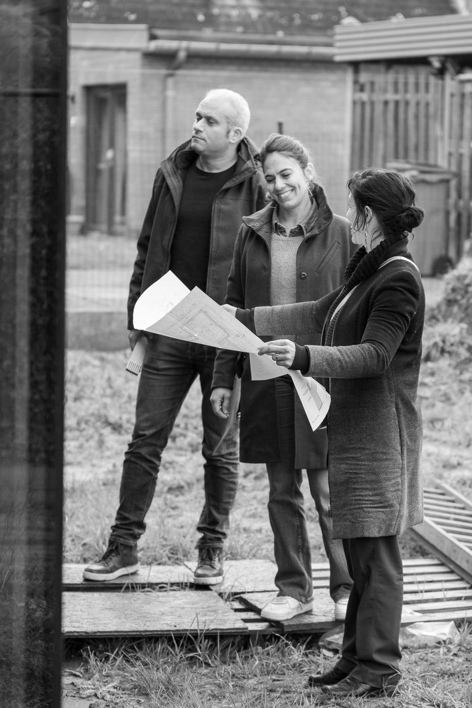 Een zwart-wit foto van drie architecten op een werf. Een vrouw houdt de plannen van een huis voor haar open.