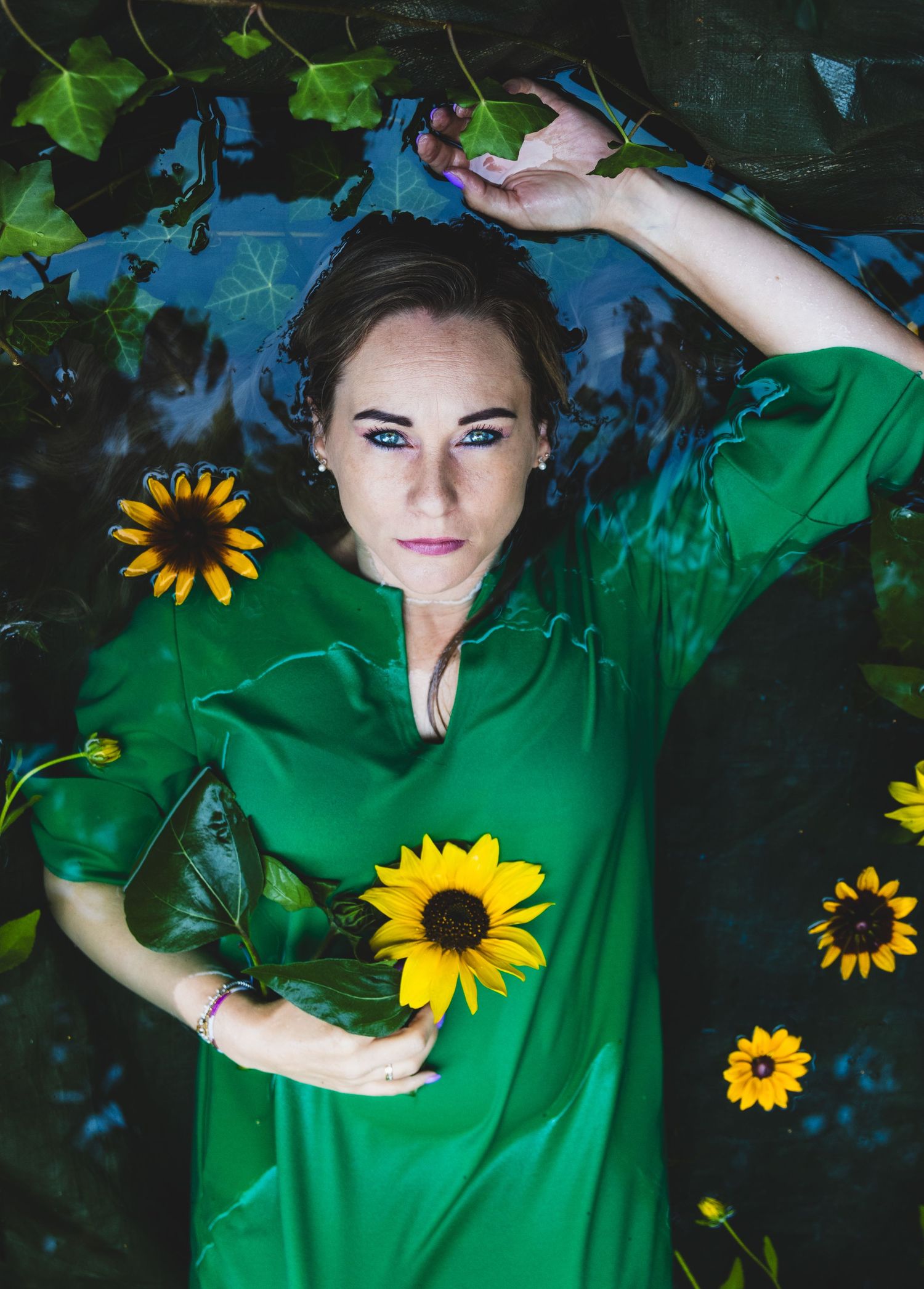 Een portret van een vrouw met doordringende blauwe ogen met een groene jurk in het water omringend met gele bloemen. Ophelia stijl
