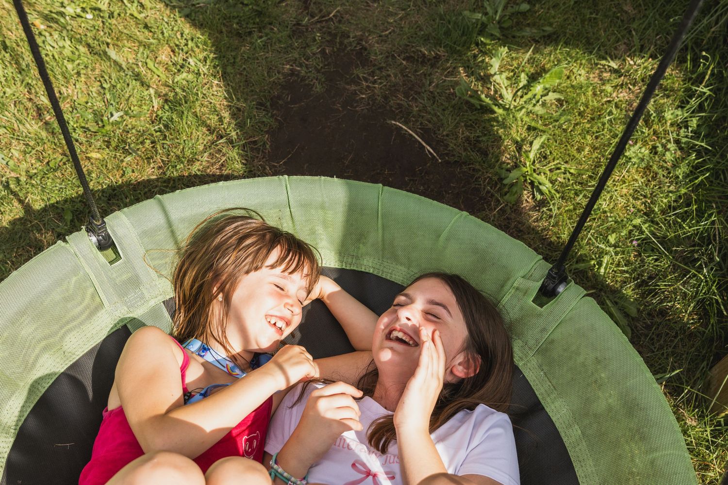 foto van twee meisjes op een ronde schommel liggen en die met elkaar aan het lachen zijn. De zon schijnt. Beeld is van bovenaf gemaakt
