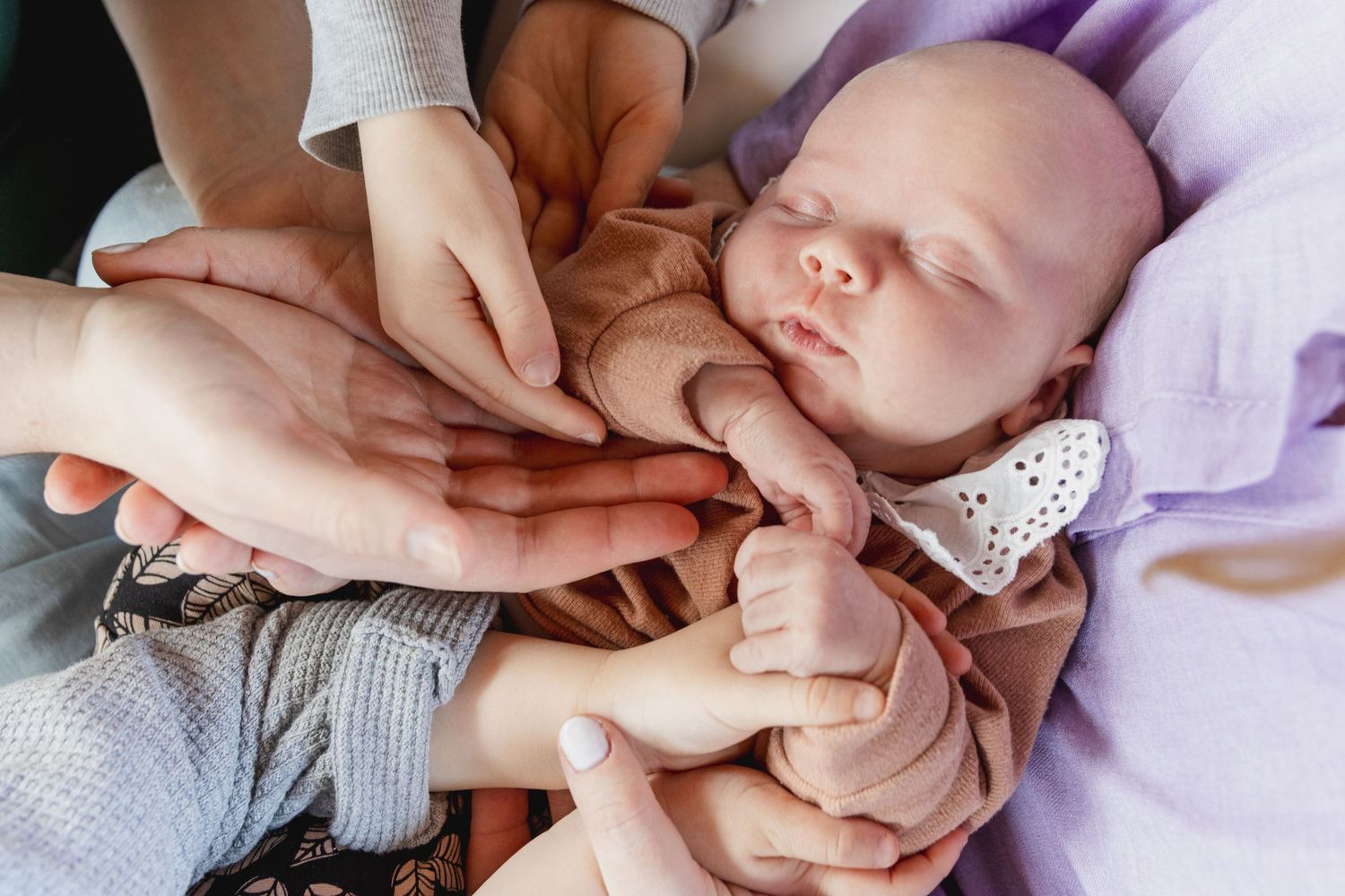 een slapende baby, omringd met de handen van vader, moeder, en broers. Ze ligt op een lavendelkleurig deken