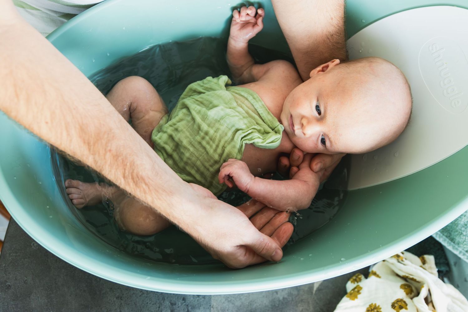 Een baby in groen badje, bedekt met een groene tetradoek. de armen van de vader die hem wassen