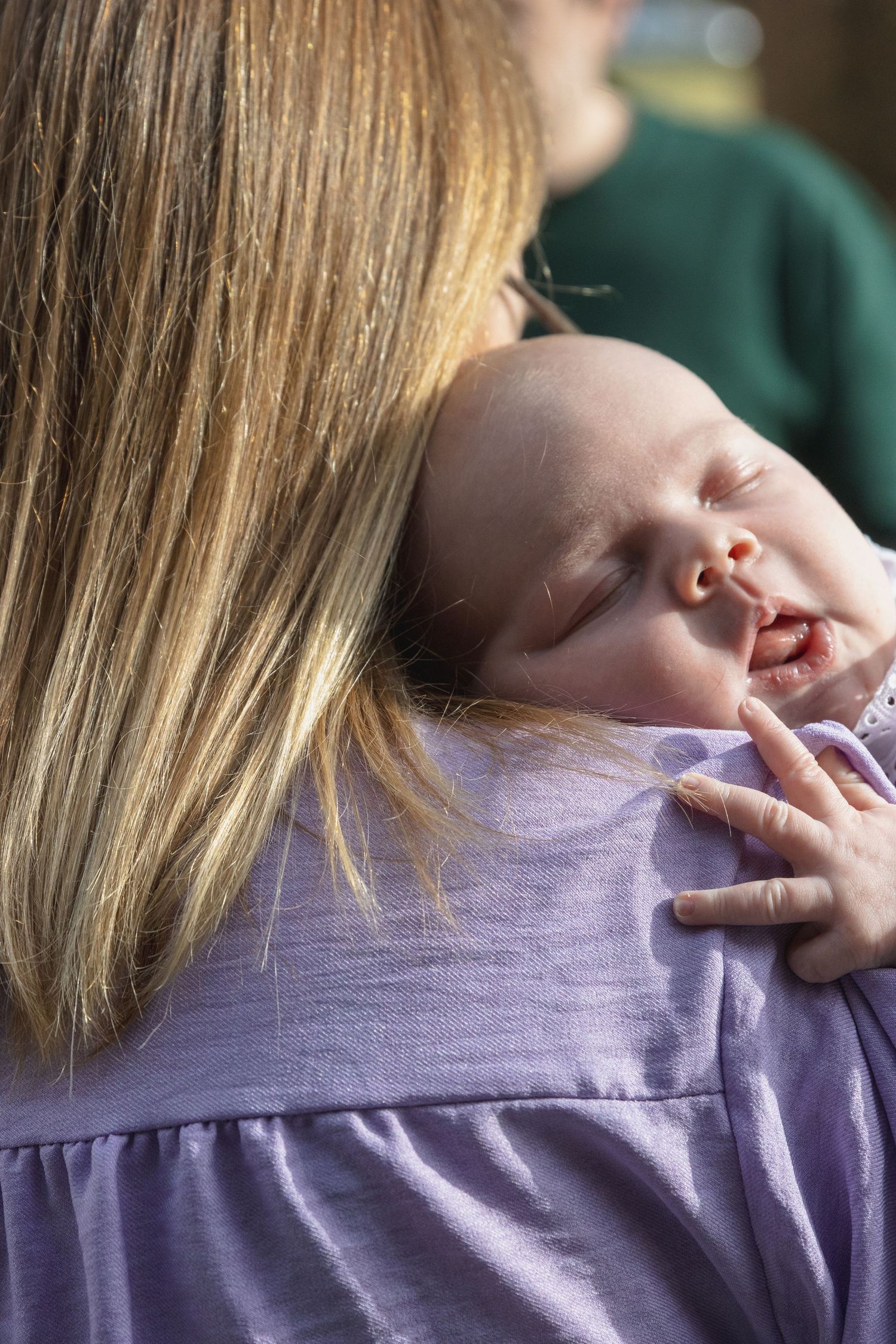 Een baby slaapt op haar moeders schouder, wegkijken van de camera. De moeder heeft stijl blond haar en draagt een lavendelkleurige trui