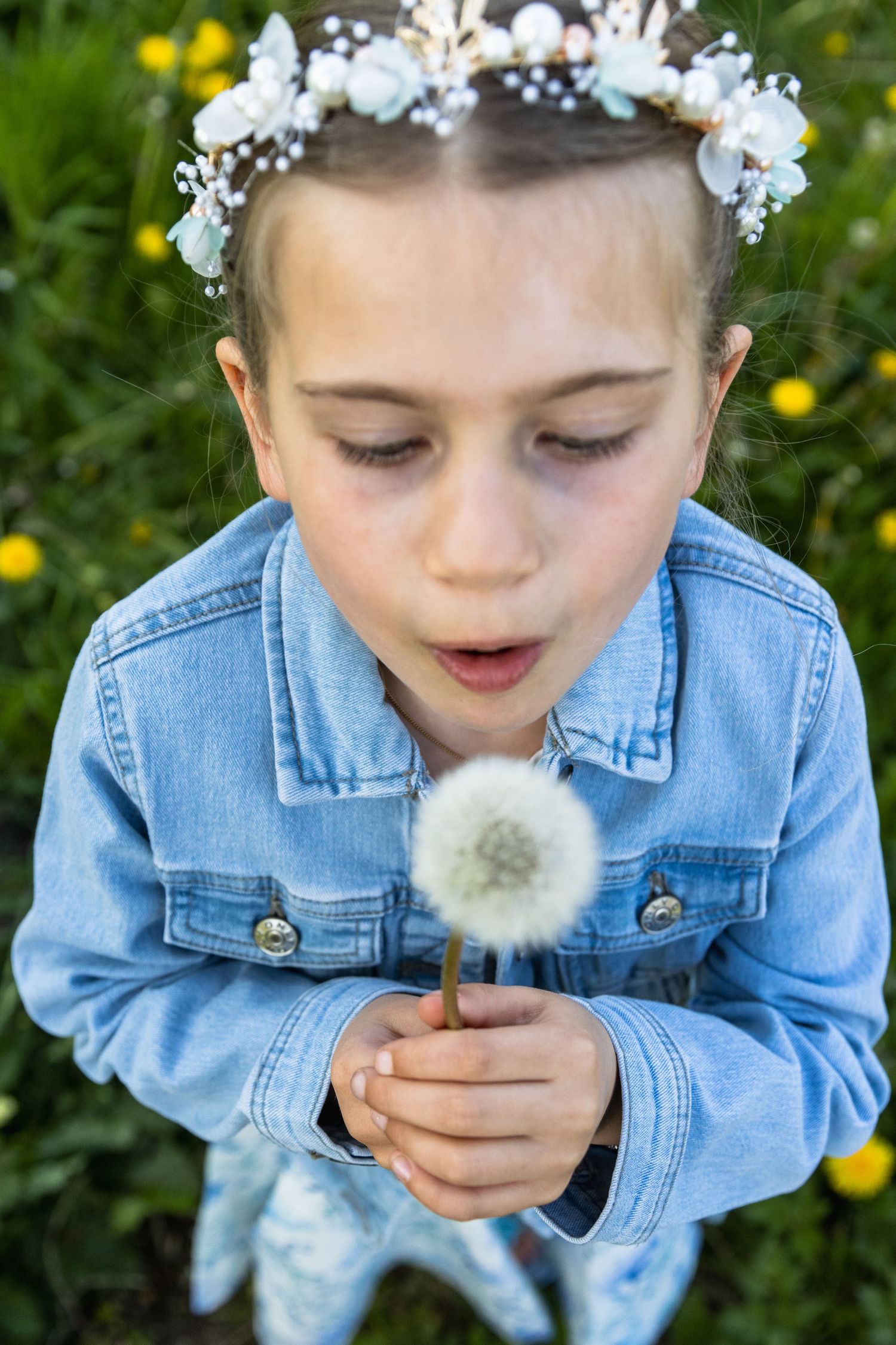 communiefoto van 7 jarig meisje die de zaadjes van een paardenbloem wegblaast. Blauw jeansjasje en bloemen in het haar