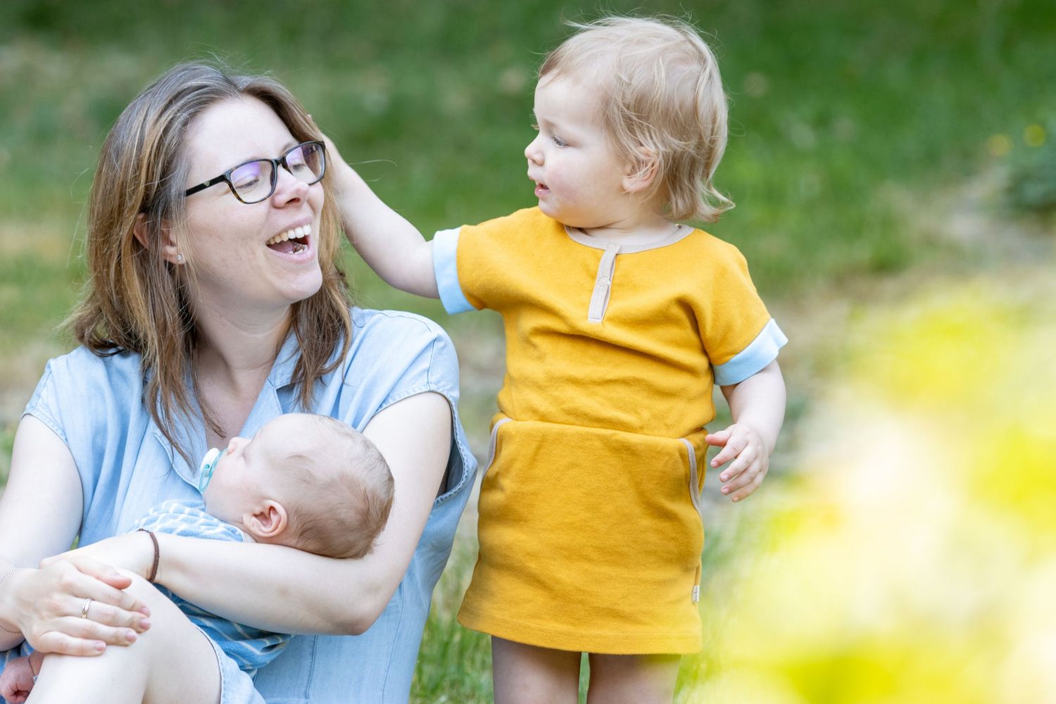 Een lachende moeder met een blauwe jurk. Haar tweejarige dochter met een gele jurk, die de moeder liefdevol streelt.  Een baby bij de moeder