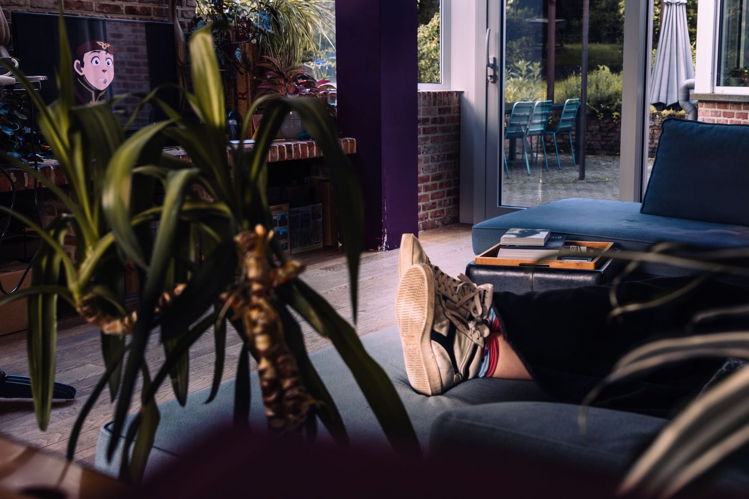 Een foto van een living met grote huisplanten. Je ziet de voeten van een vrouw. Ze draagt diadoras en op de achtergrond een tv met avatar.