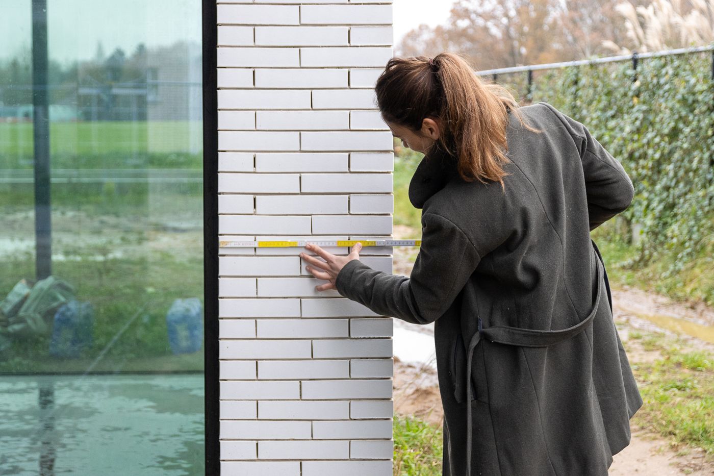 Een architecte gericht met haar rug naar de lens. Ze meet met een gele meter de witte stenen muur op.