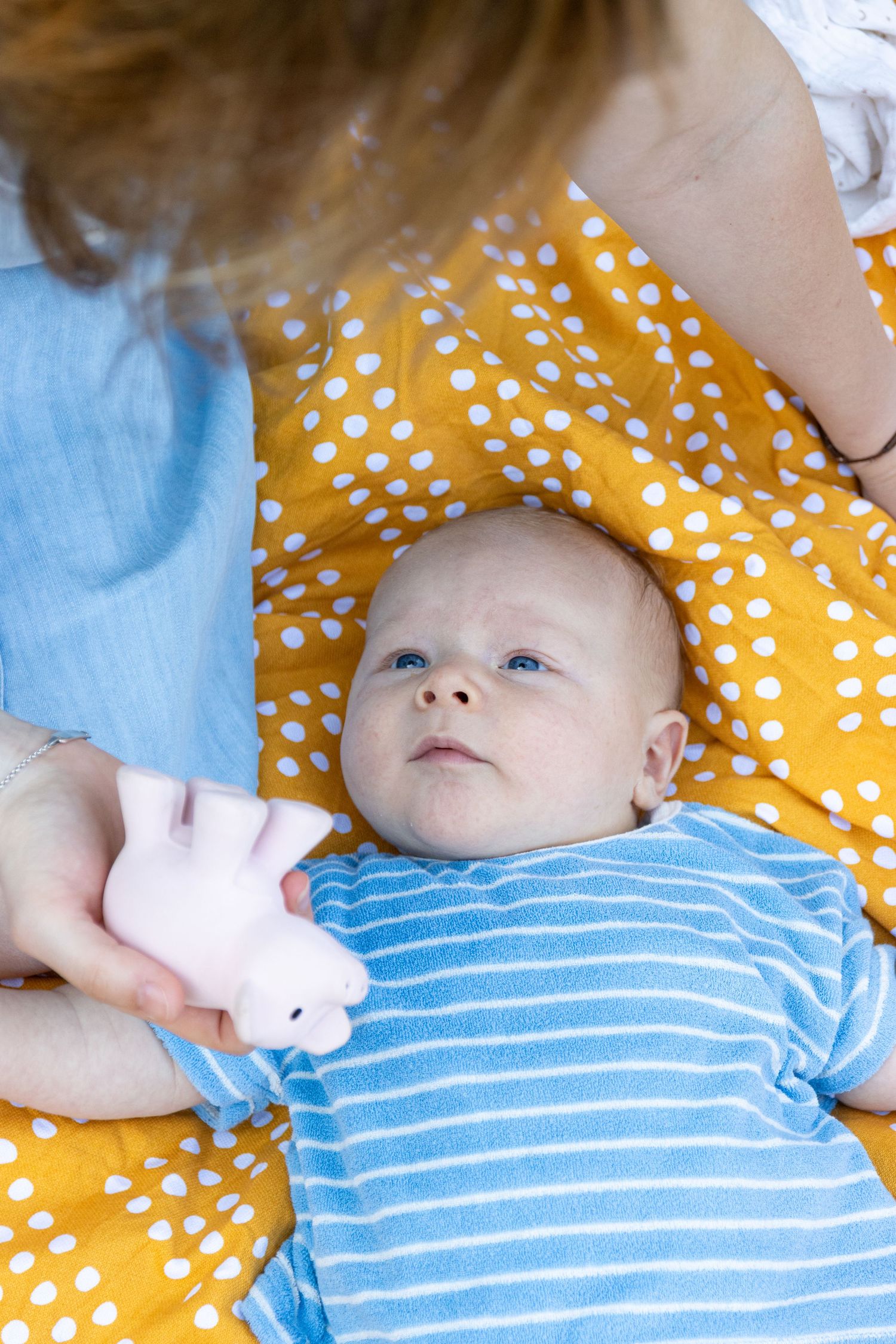 een baby met een blauw gestreept t-shirt ligt op zijn rug op een geel deken met witte stippen. een vrouw geeft een speelgoedbeer.