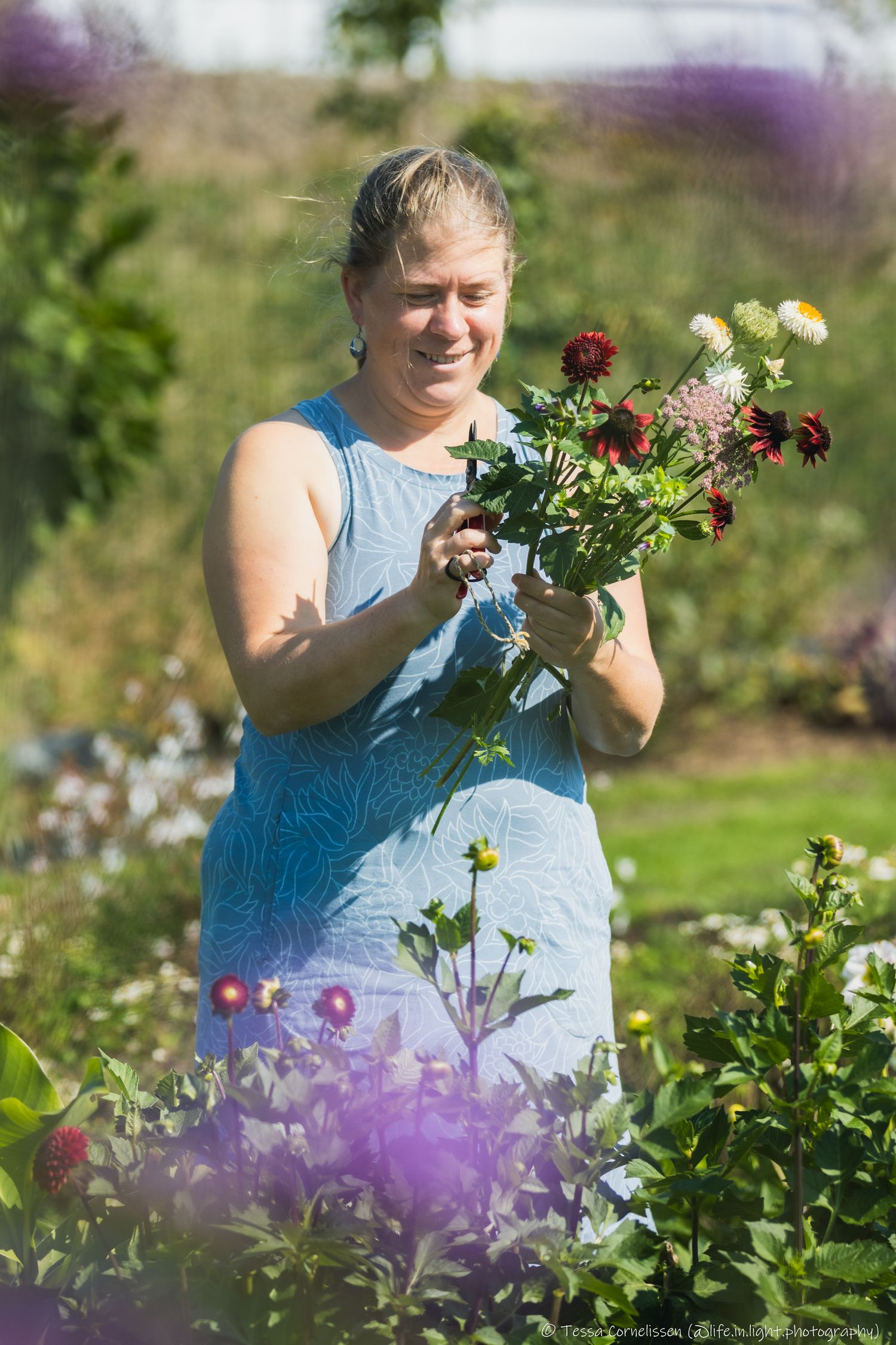 Een portret van een lachende vrouw in een blauwe jurk. Ze staat in een pluktuin en houdt een boeket vast met vers geplukte bloemen.