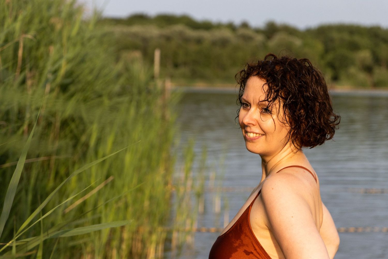Portret van een glimlachende vrouw. Vanaf de borst, halflange krullende haren. Ze draagt een bruin zwempak en staat in het water tussen riet