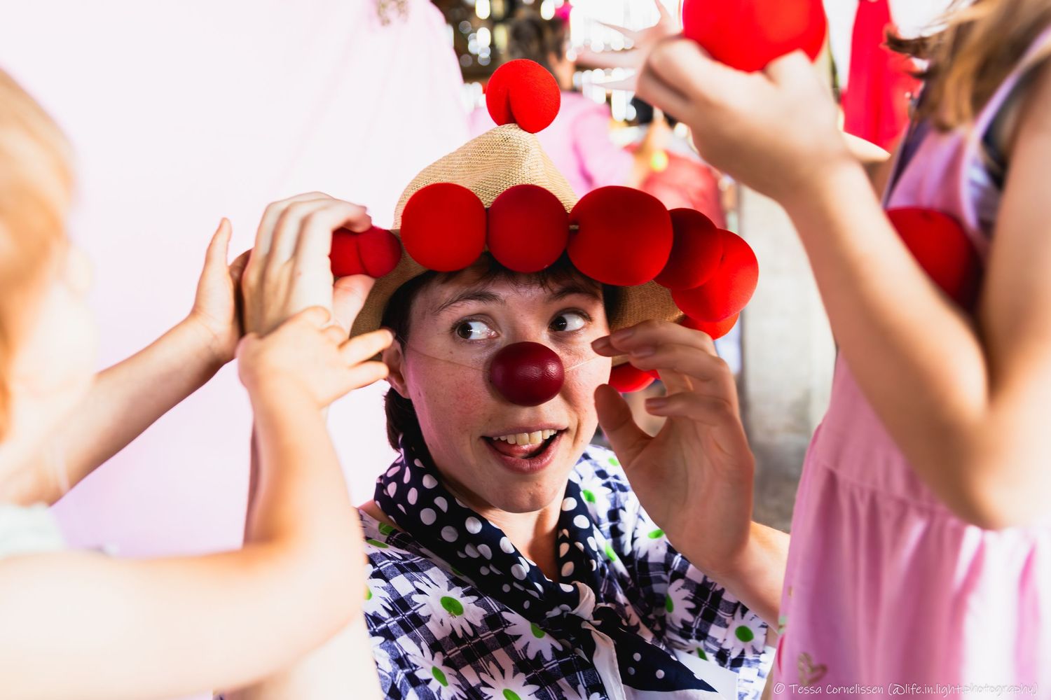 cliniclowns. Een vrouw met een grote hoed waarop twee meisjes die je maar gedeeltelijk ziet grote rode ballen plaatsen.