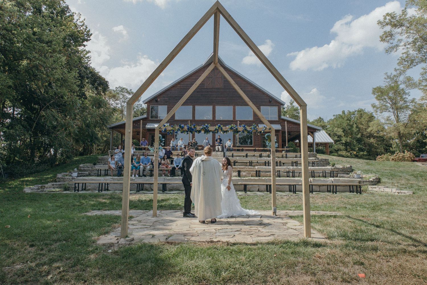 Wedding ceremony taking place in a rustic wooden chapel structure with guests seated on benches under blue skies.