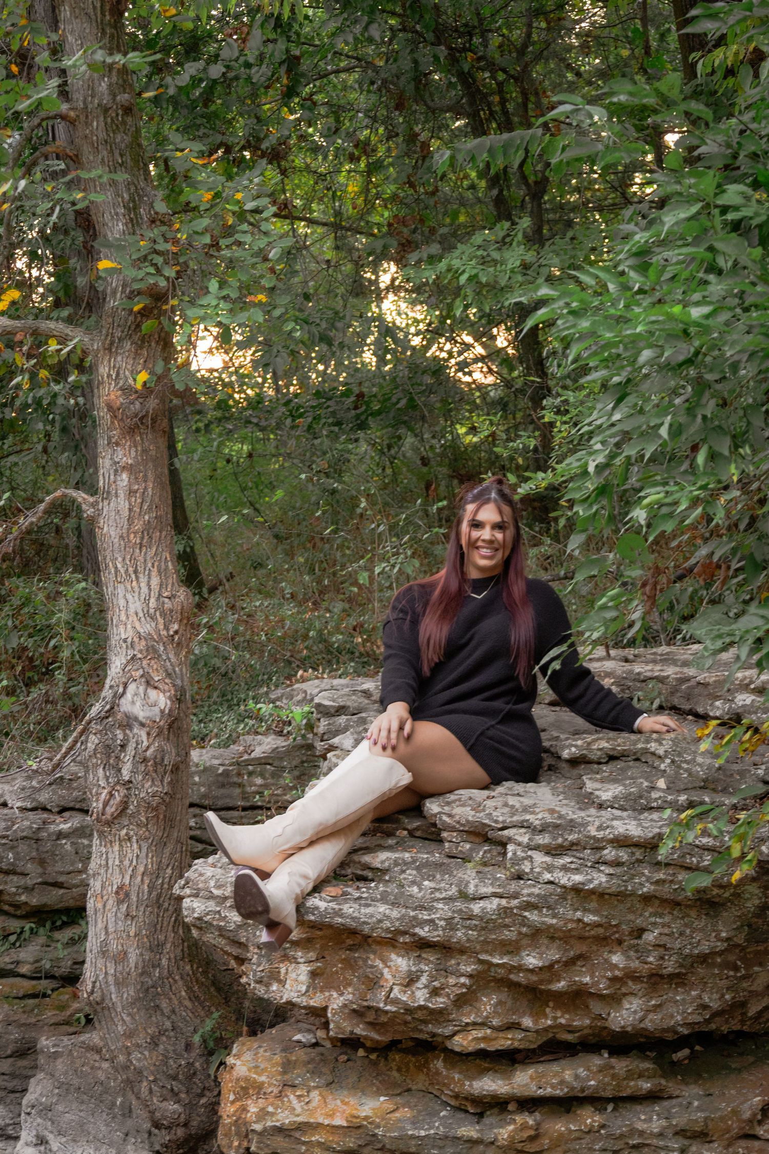 Person in black clothing sitting on a rocky ledge in a wooded area showing off their leg cast.