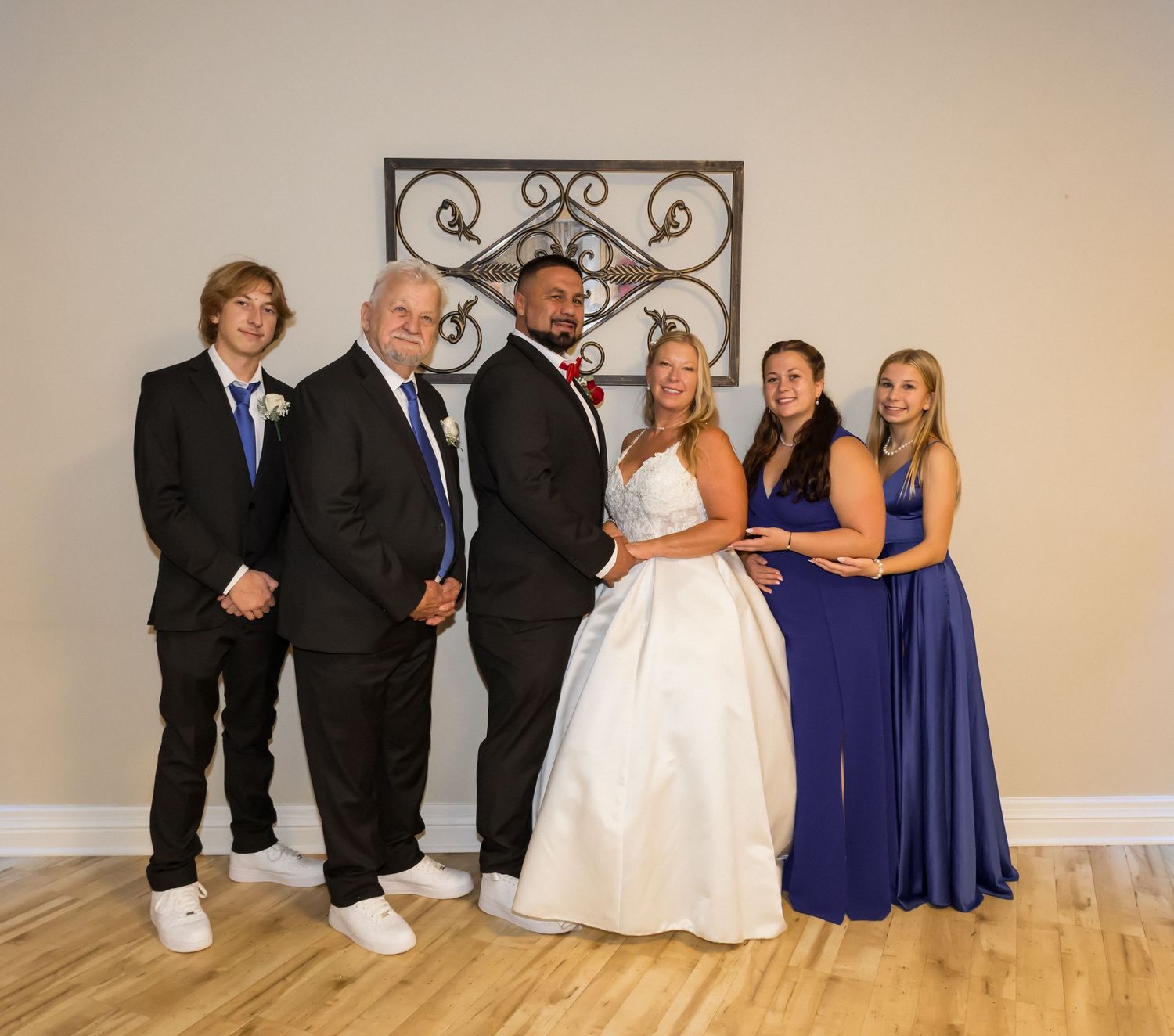 Wedding party in formal attire poses against light colored wall with bride in white gown and bridesmaids in blue dresses.