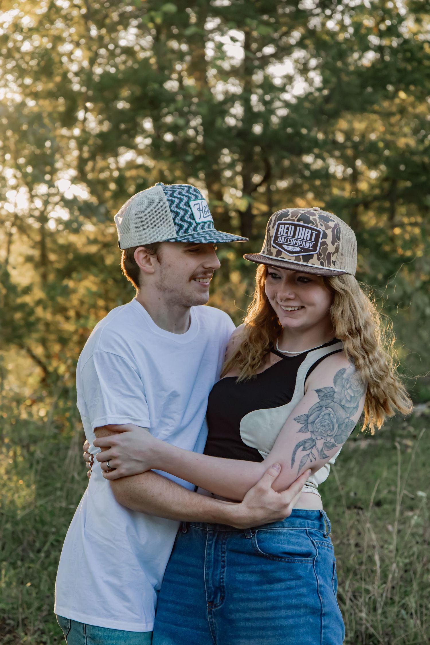 A couple shares a tender moment while wearing casual attire against a backdrop of trees and sunlight.