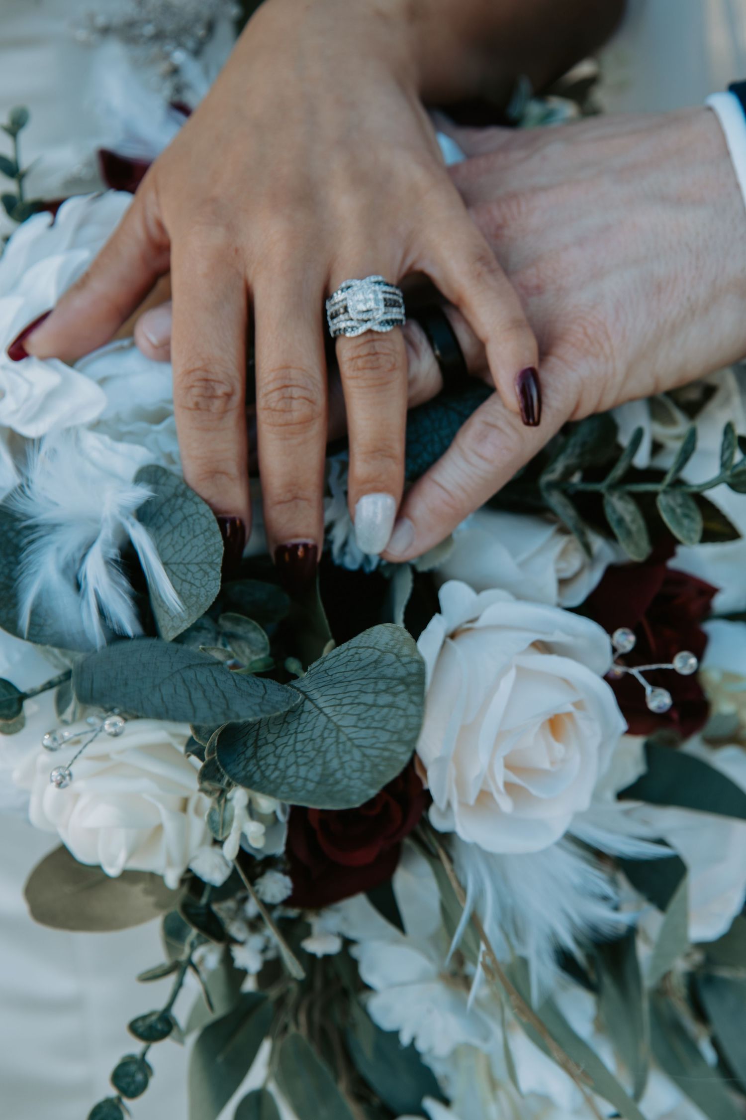 Hand wearing a sparkling engagement ring rests on elegant bouquet of white roses and burgundy flowers with eucalyptus leaves.