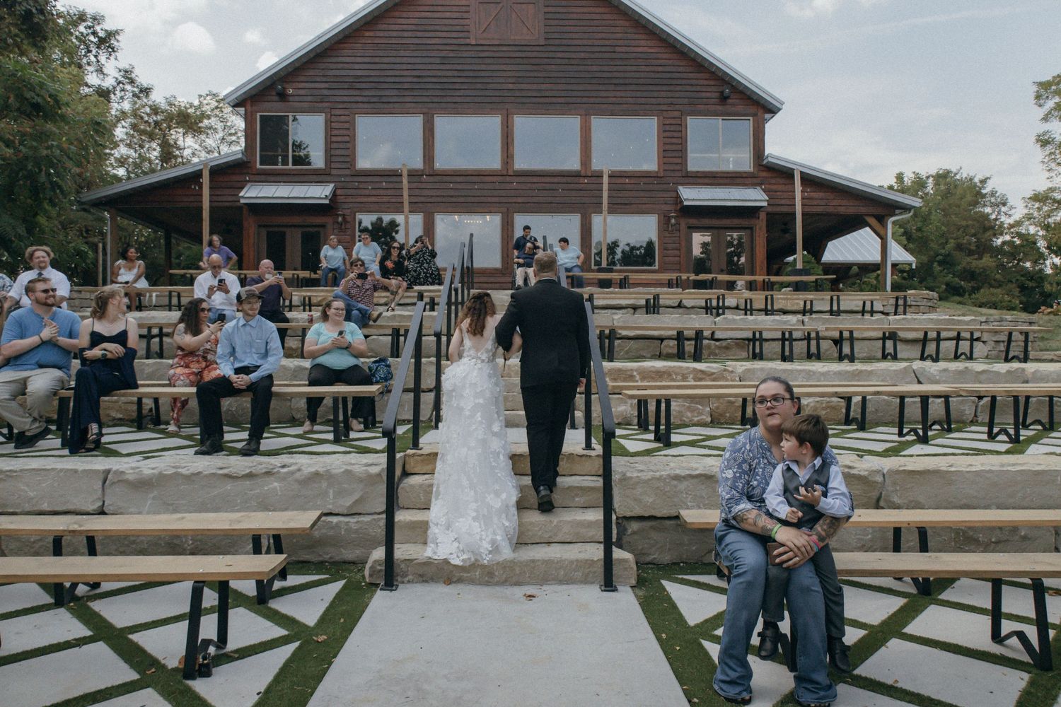 A rustic outdoor wedding venue with wooden benches and a large lodge-style building in the background.