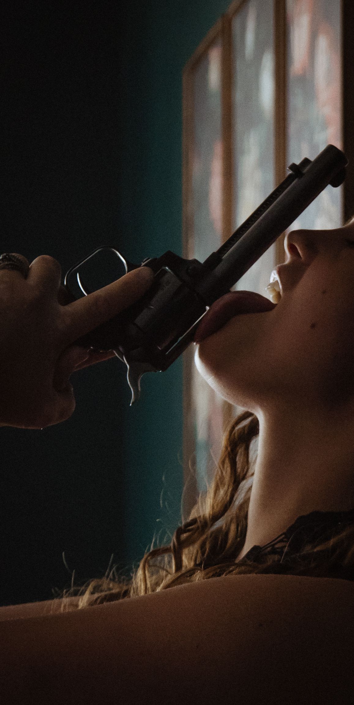A dark silhouette of a person lying down with a handgun held against their face in dramatic lighting.