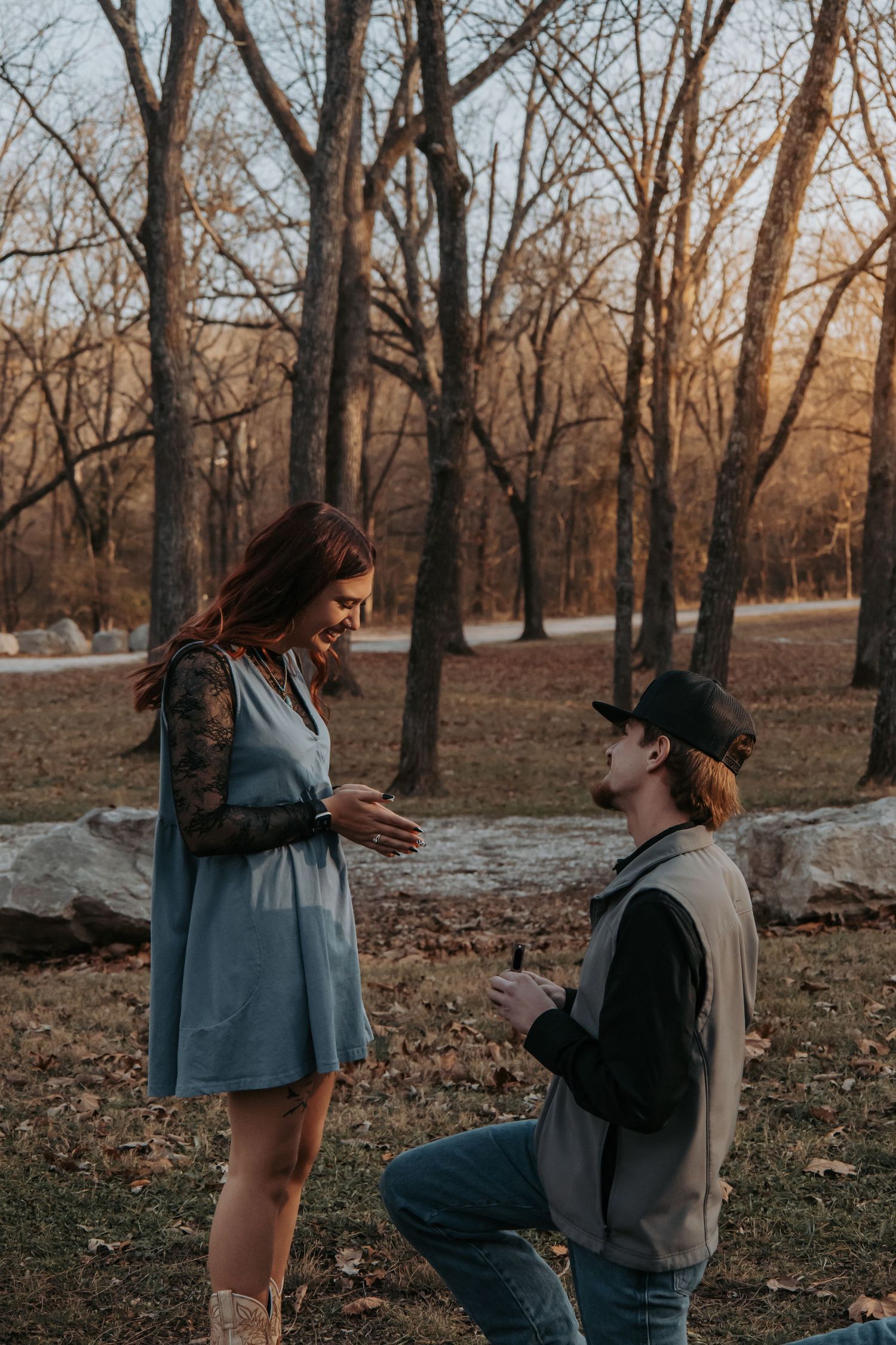 A couple shares a tender moment in a wooded park during autumn with bare trees in the background.