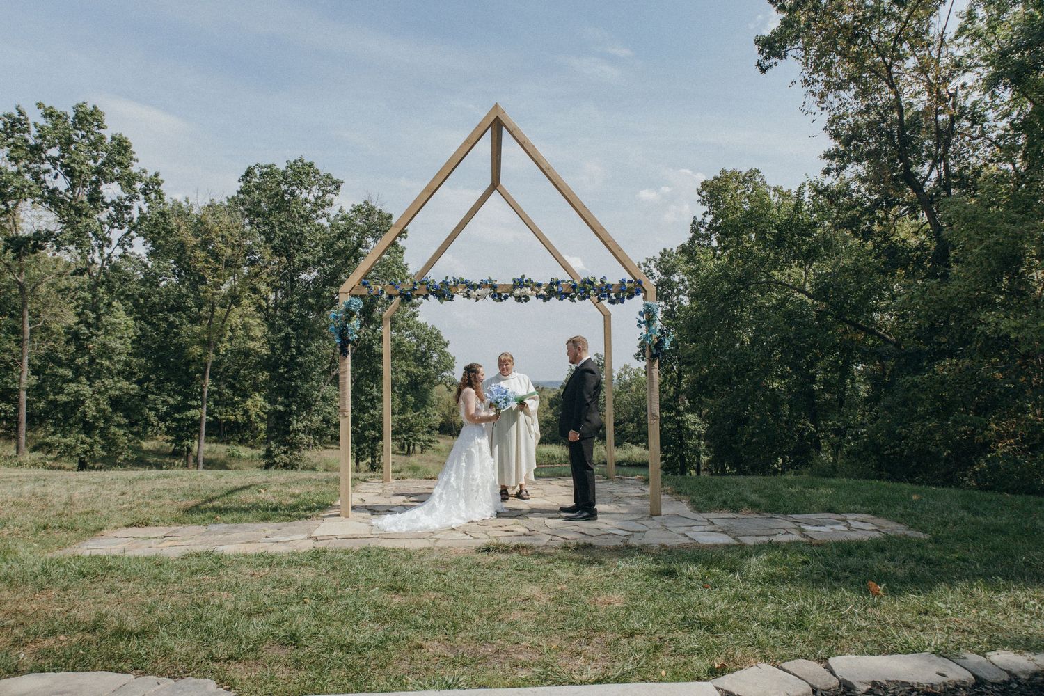 A triangular wooden wedding arch stands in a rustic outdoor setting with trees in the background.