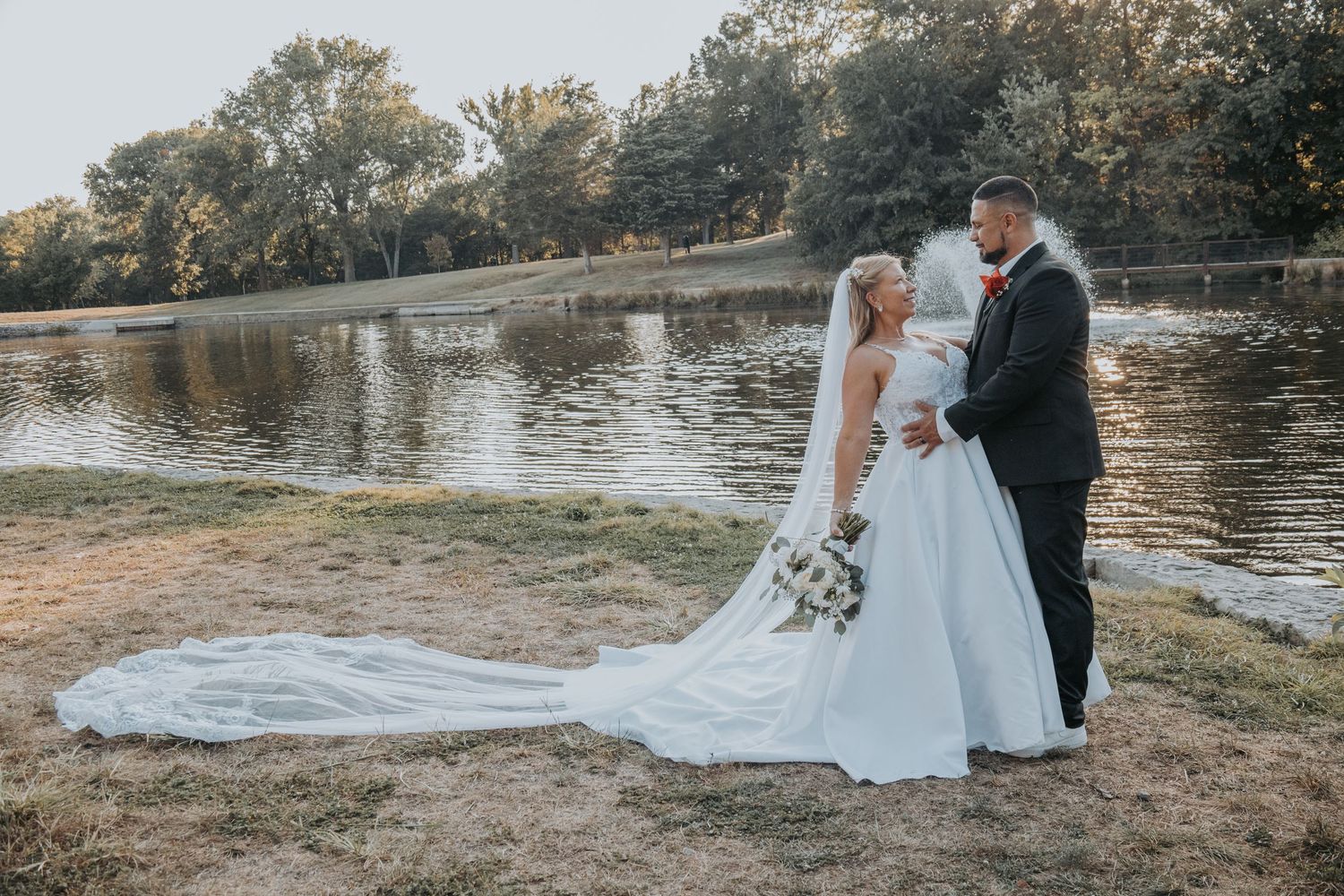 A couple in wedding attire shares romantic moments by a serene lake during their outdoor autumn wedding photoshoot.