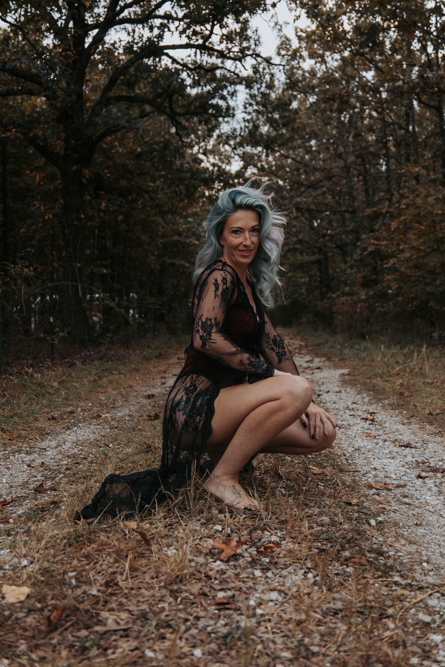 A woman in a black dress sits on a gravel path surrounded by autumn foliage in a moody forest setting.