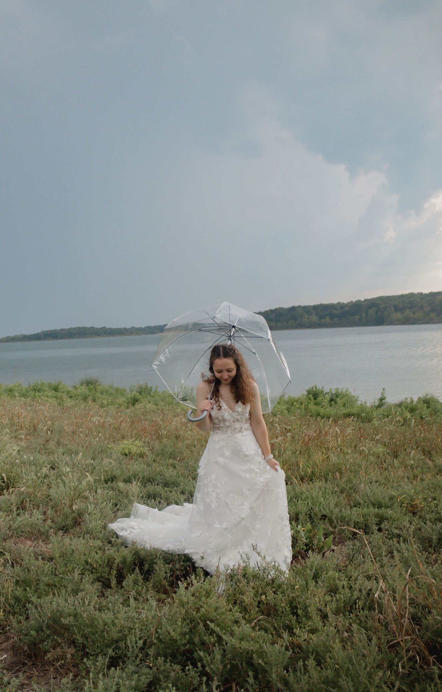 A flowing white wedding dress twirls gracefully against a scenic coastal landscape with grassy hills and water views.